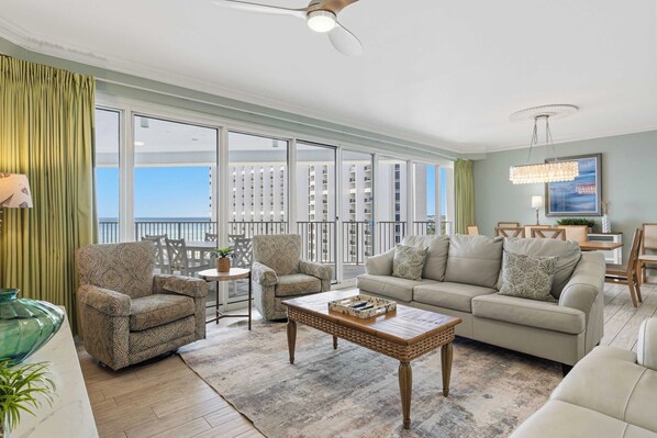 A spacious living room with a balcony, a sofa set, a coffee table, and a view of high-rise buildings. - Beachfront escape with Gulf views, pool access (Miramar Beach)