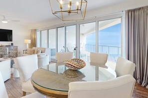 Moderni hotellihuone, jossa on lasinen ruokapöytä, valkoiset tuolit, koristeellinen kulho, taulutelevisio ja parveke, jolta on näkymä.