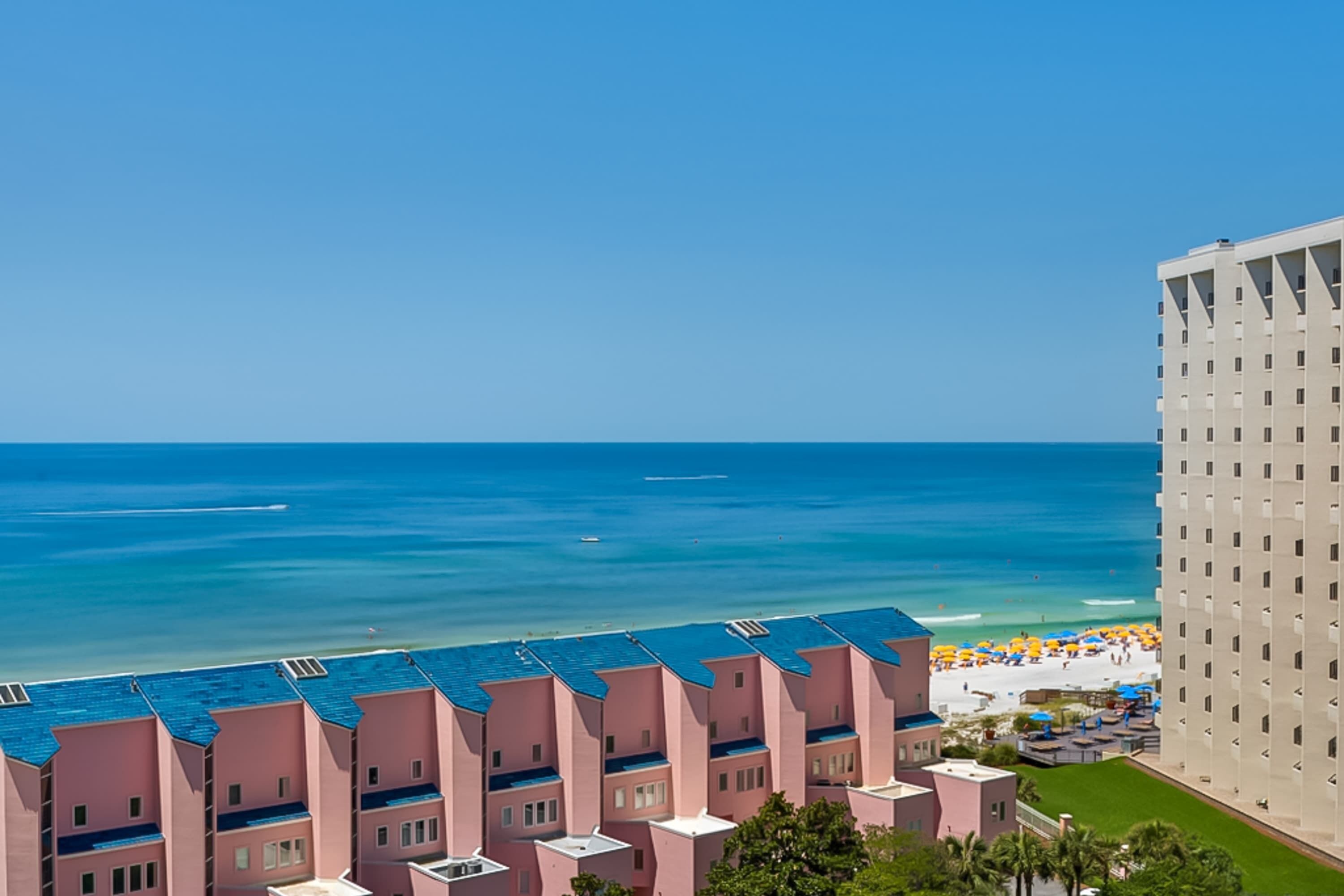 A coastal hotel with multiple pink buildings, a beach with umbrellas, and a clear view of the ocean.