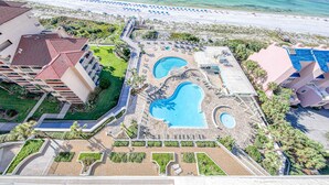 An aerial view of a resort with a central swimming area, multiple pools, and a beach with umbrellas. - Oceanfront condo with breathtaking views (Miramar Beach)