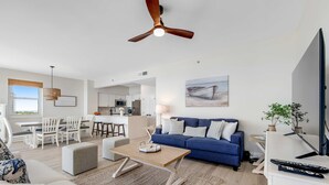 Un salon moderne avec un canapé bleu, des fauteuils blancs, une table basse en bois et un ventilateur de plafond.