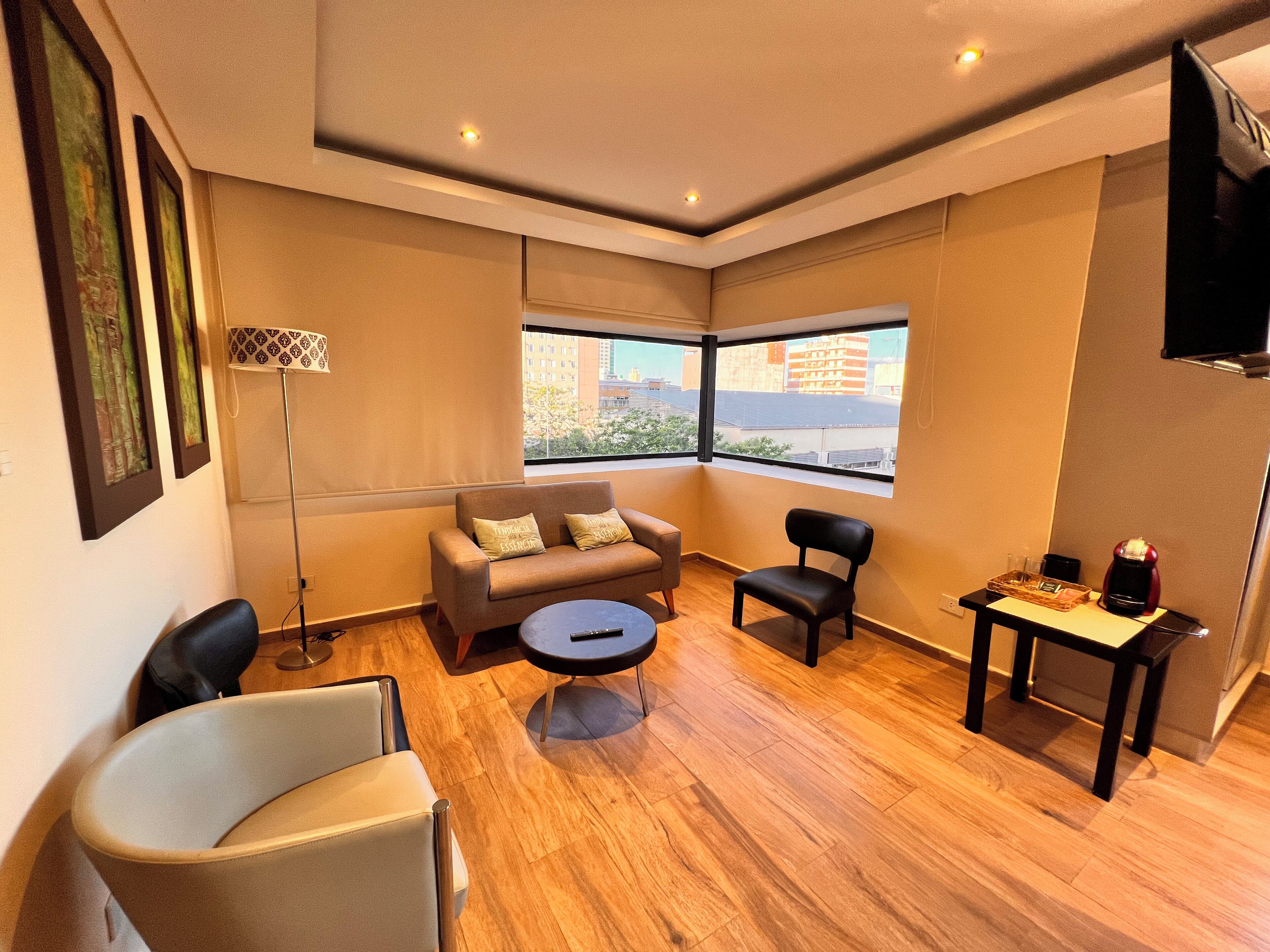 A modern living room with a sofa, chairs, a coffee table, and a small table with a coffee machine. Large windows offer a view of the city.