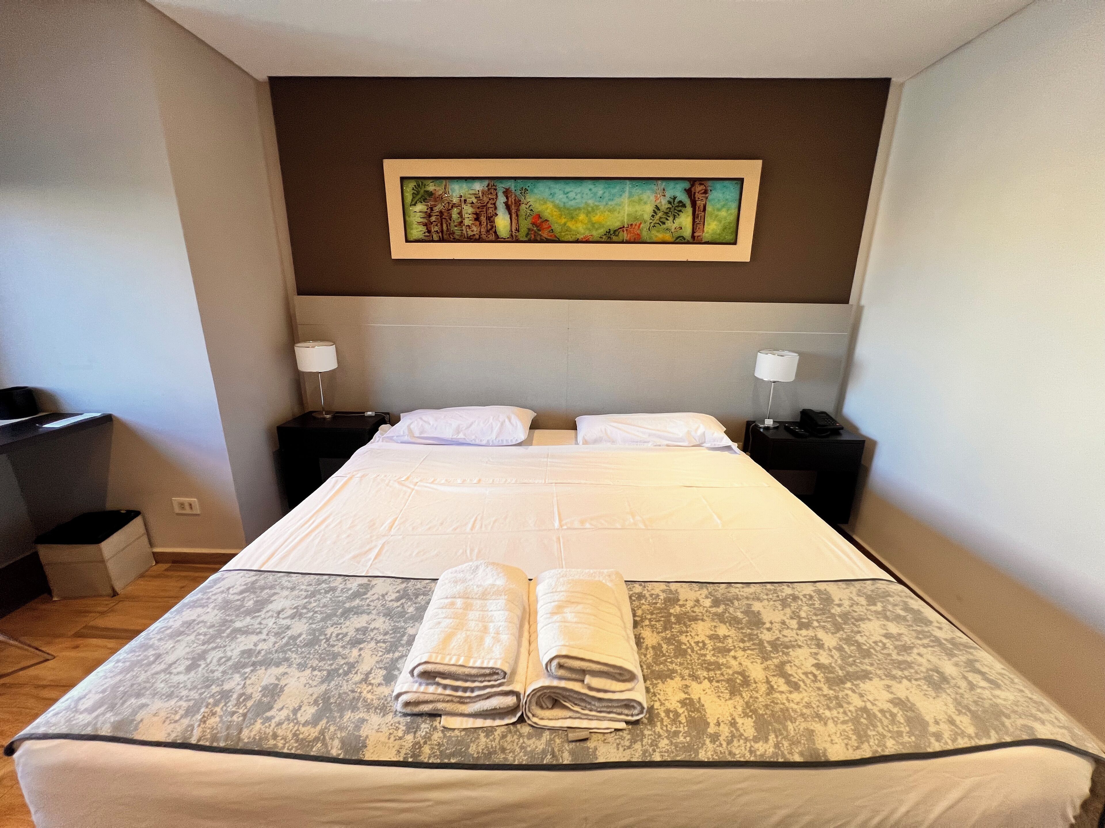 A hotel room with a large bed, two bedside tables with lamps, a painting on the wall, and a desk with a chair.