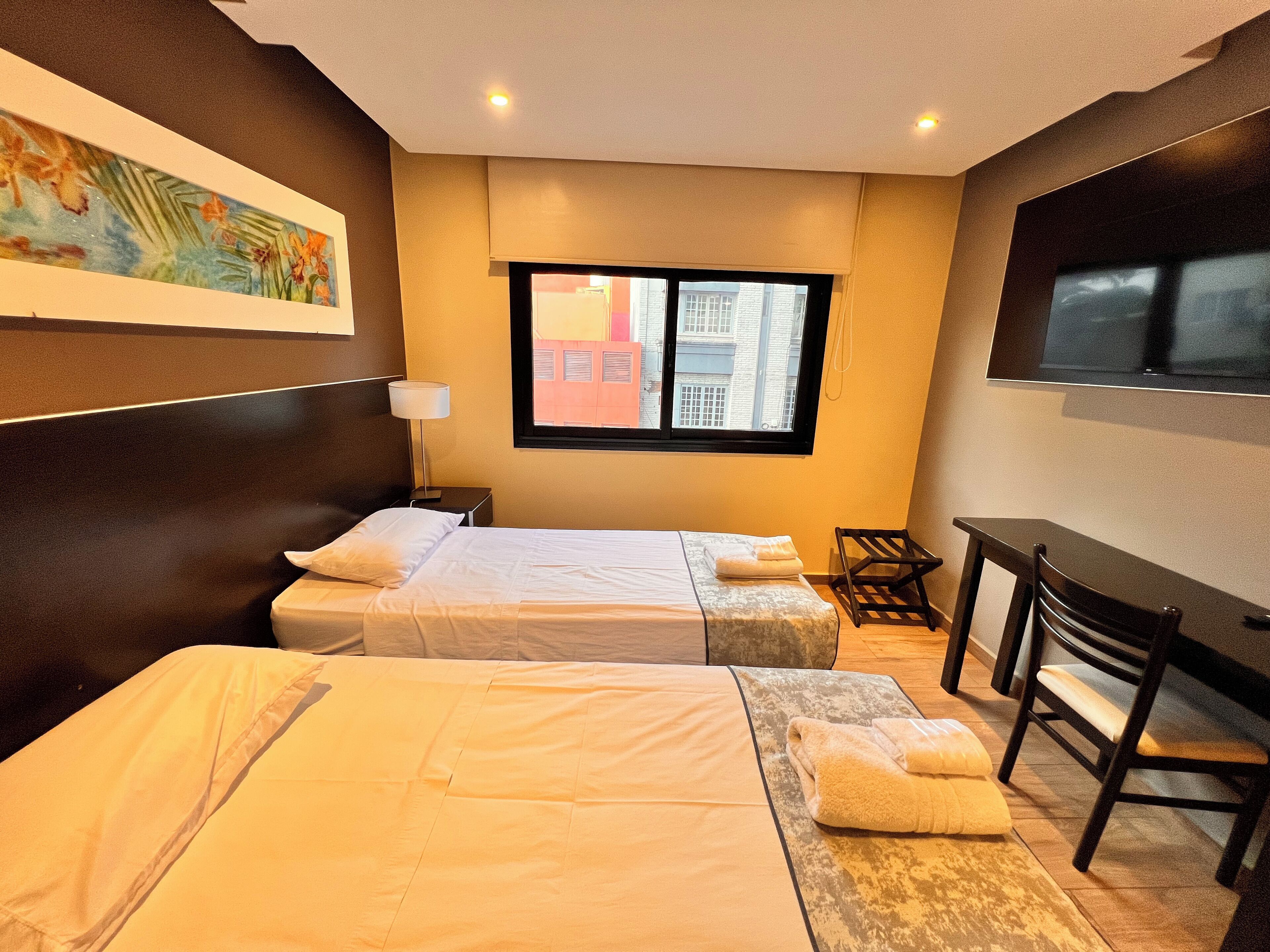A hotel room with two beds, a desk, a chair, a TV, and a window with a view of buildings.
