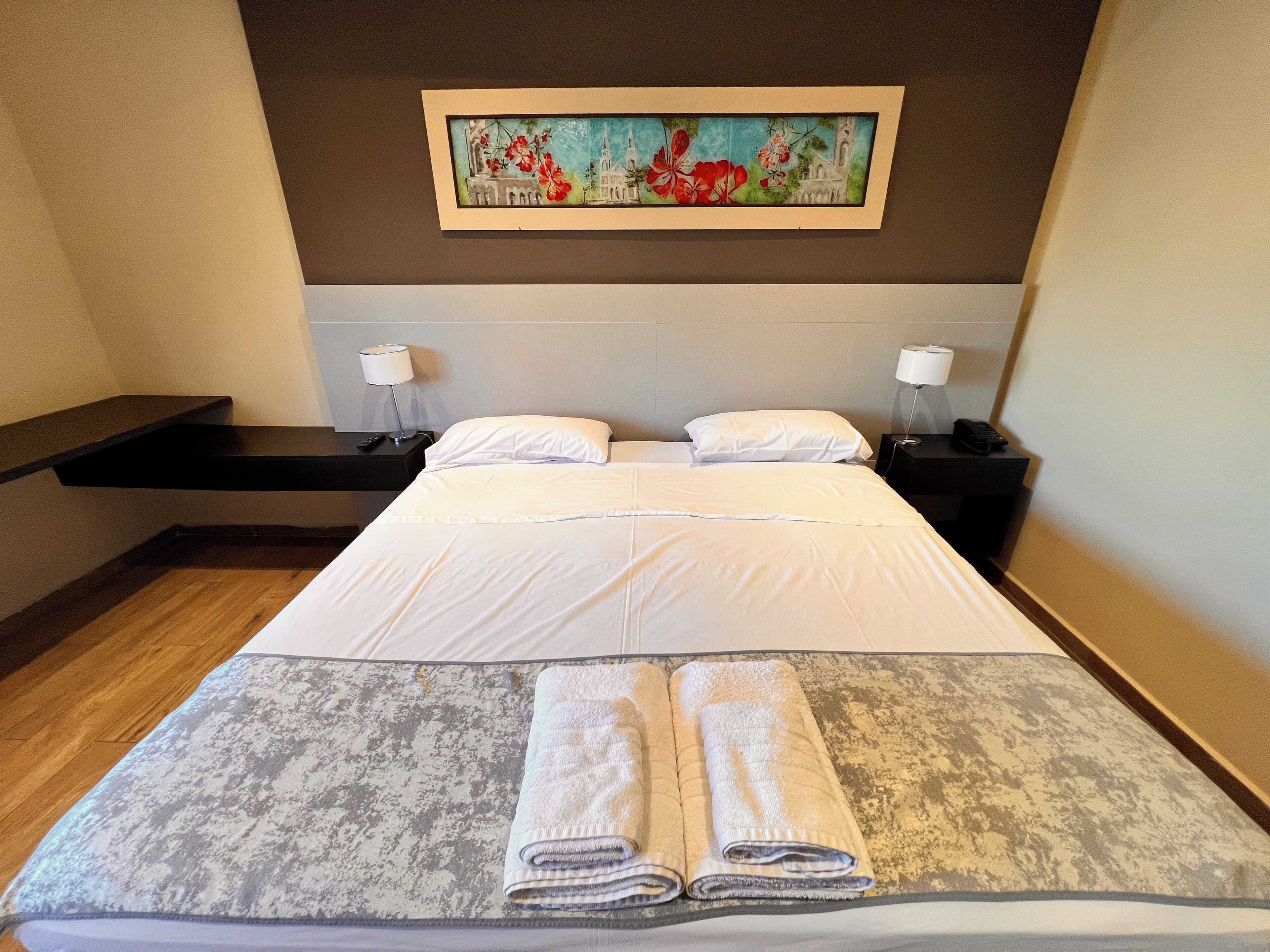 A hotel room with a bed, two bedside tables, a painting on the wall, and a wooden floor.