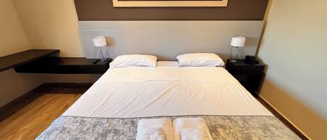 A hotel room with a bed, two bedside tables, a painting on the wall, and a wooden floor.