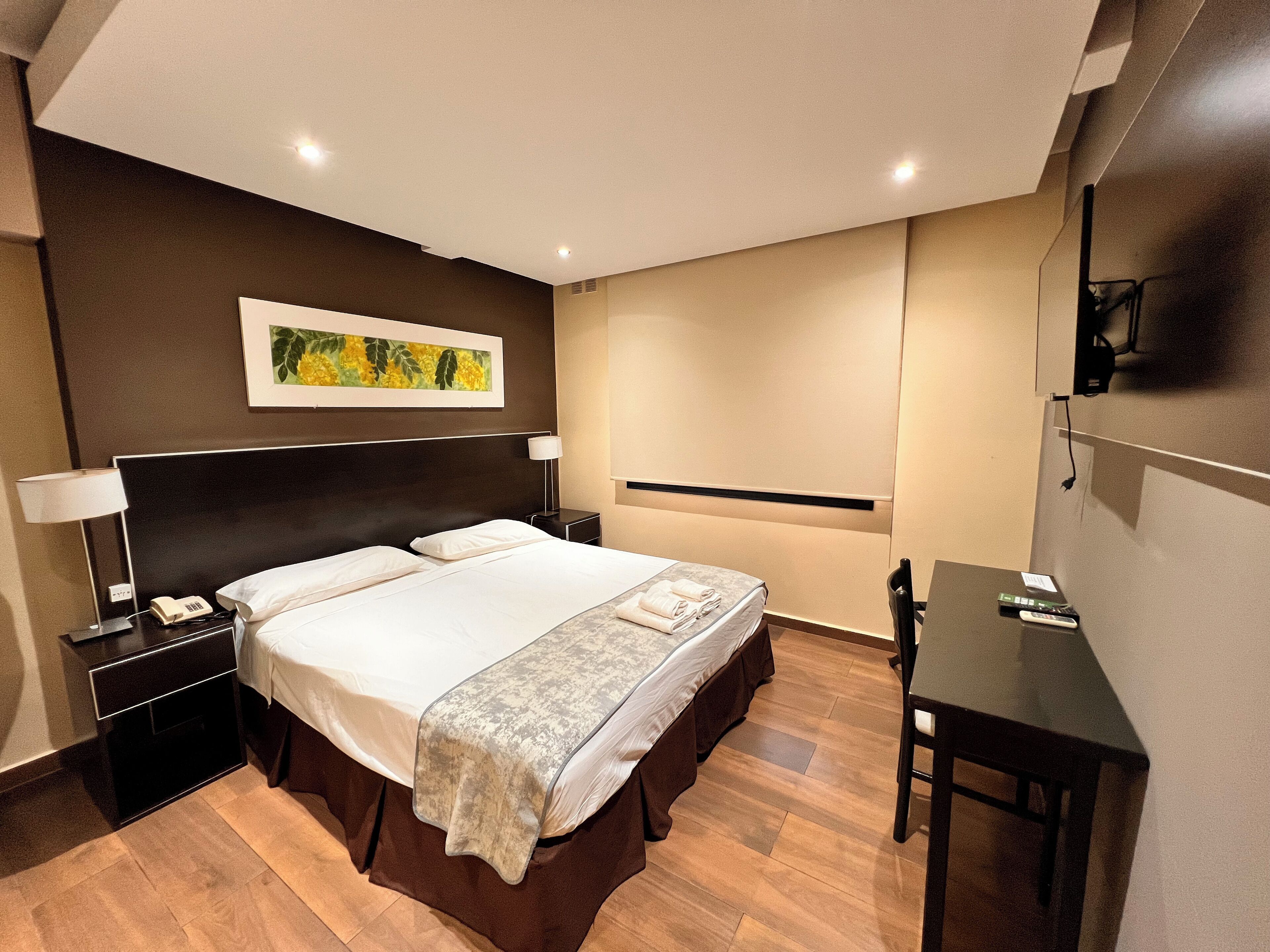 A modern hotel room with a large bed, bedside tables, a desk, and a painting on the wall.