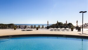 Un área de piscina con sillones de descanso y sombrillas, con vista despejada a la playa y un puesto de salvavidas.