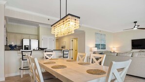 Un comedor con una mesa larga, sillas blancas y un candelabro. Se observa una cocina al fondo con armarios blancos y un refrigerador.