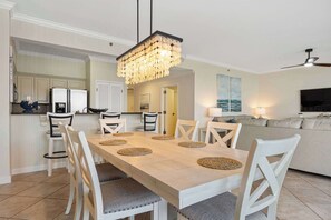 A dining area with a long table, white chairs, and a chandelier. There is a kitchen in the background with white cabinets and a refrigerator. - Steps to Beach: Ocean View, Hot Tub + Pools (Miramar Beach)