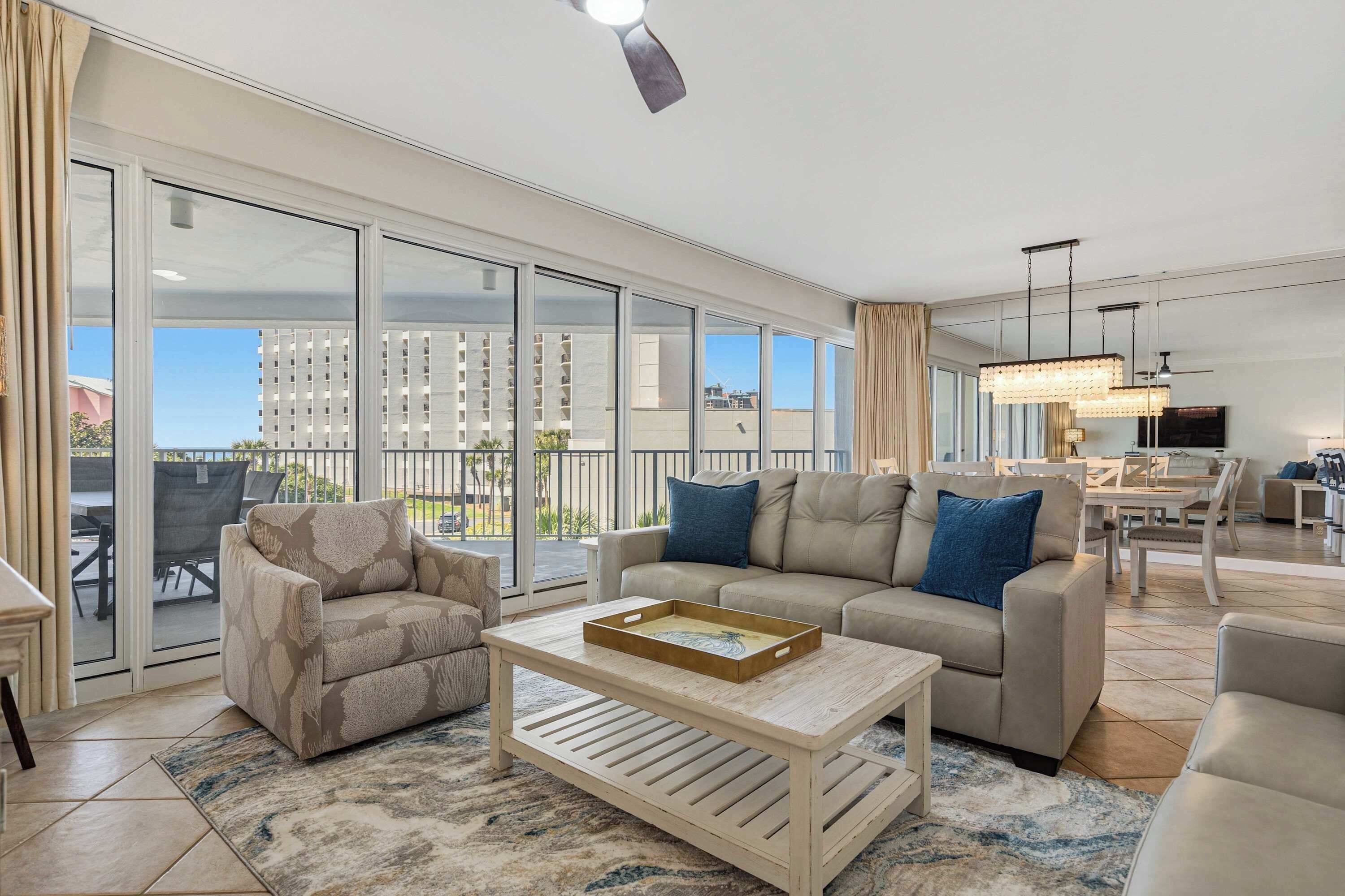 A spacious living area with a sofa, armchair, and coffee table, large sliding glass doors, and a view of buildings.