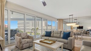 Amplia sala de estar con sofá, sillón y mesa de centro, puertas correderas de cristal y vistas a edificios.