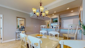 A dining area with a glass table, white chairs, a chandelier, and a kitchen in the background. - Exclusive 2BR Beachfront | Gulf Views Balcony (Miramar Beach)