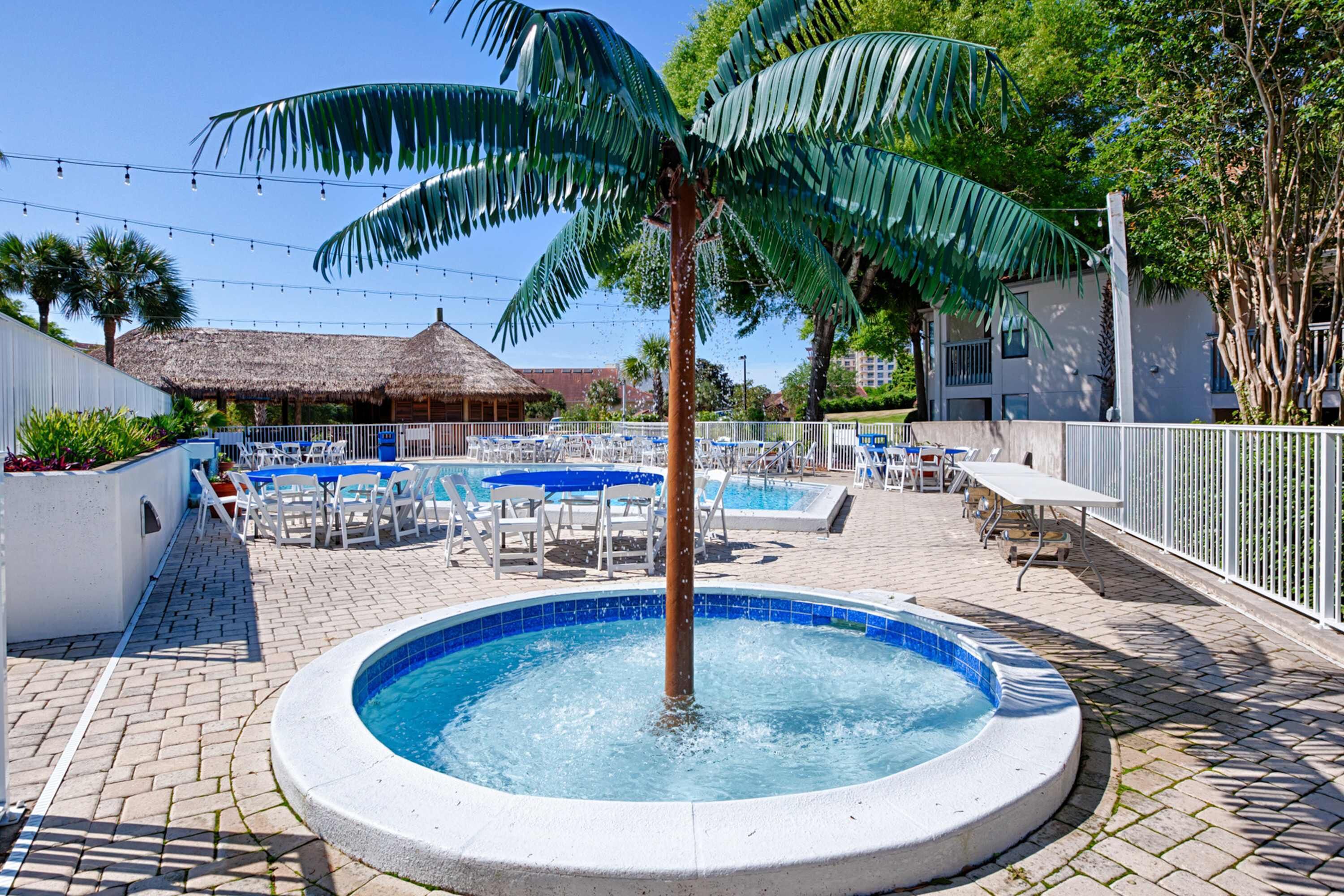 Een zwembadgedeelte met een centraal gelegen hottub, omringd door ligbedden en tafels, palmbomen en een gebouw met rieten dak op de achtergrond.