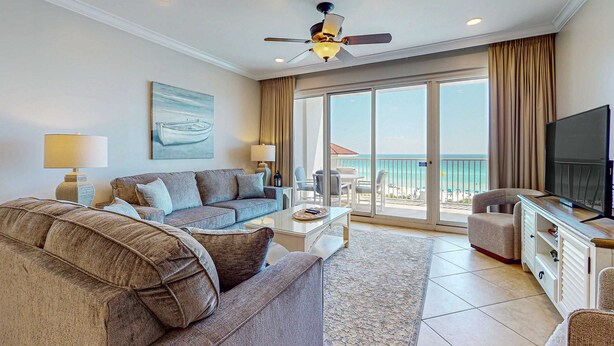 A spacious living room with a sofa, coffee table, and TV stand, a ceiling fan, and a large sliding glass door leading to a balcony with a view of the sea. - Exclusive 2BR Beachfront | Gulf Views Balcony (Miramar Beach)