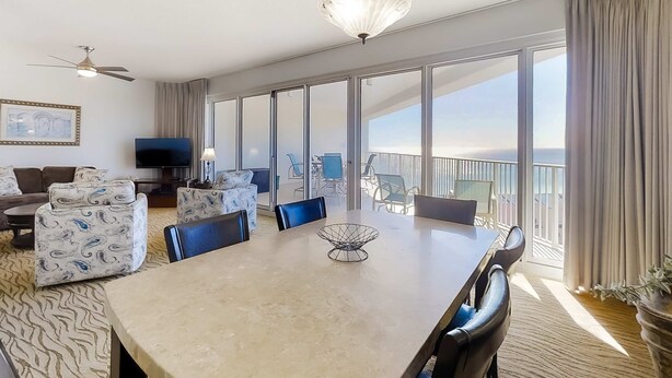 Un vaste espace de vie comprenant une grande table à manger, des sièges confortables, un ventilateur de plafond et un balcon avec vue.