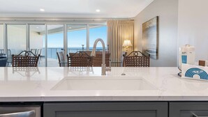 Una cocina moderna con una isla grande, sillas de mimbre, vistas al mar y un cuadro en la pared.