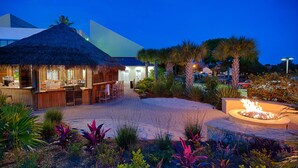 A thatched-roof outdoor dining area with a fire pit, surrounded by tropical plants and a modern building in the background. - 2BR Gulf-front condo with ocean views (Miramar Beach)