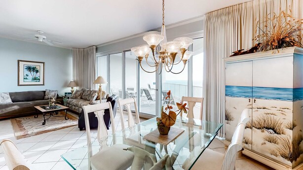 A modern living room with a glass dining table, a chandelier, and a view of the sea through large windows. - 2BR Gulf-front condo with ocean views (Miramar Beach)