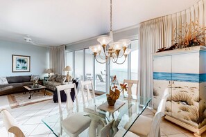 Una sala de estar moderna con una mesa de comedor de cristal, una araña y vistas al mar a través de amplios ventanales.