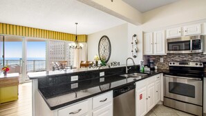 Cozinha moderna com armários brancos, bancada de granito preto, eletrodomésticos de aço inoxidável e vista para o oceano.