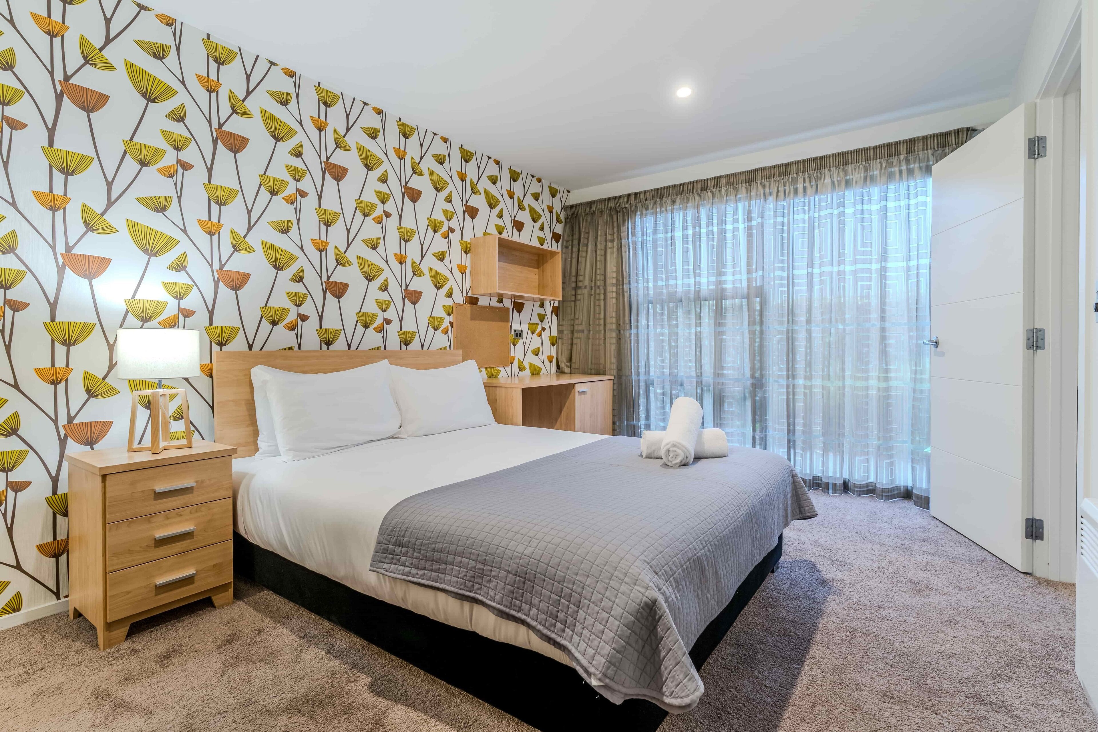 A neatly arranged bedroom with a large bed, a bedside table with a lamp, a nightstand, a wall-mounted shelf, and a window with curtains.
