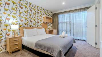 A neatly arranged bedroom with a large bed, a bedside table with a lamp, a nightstand, a wall-mounted shelf, and a window with curtains.