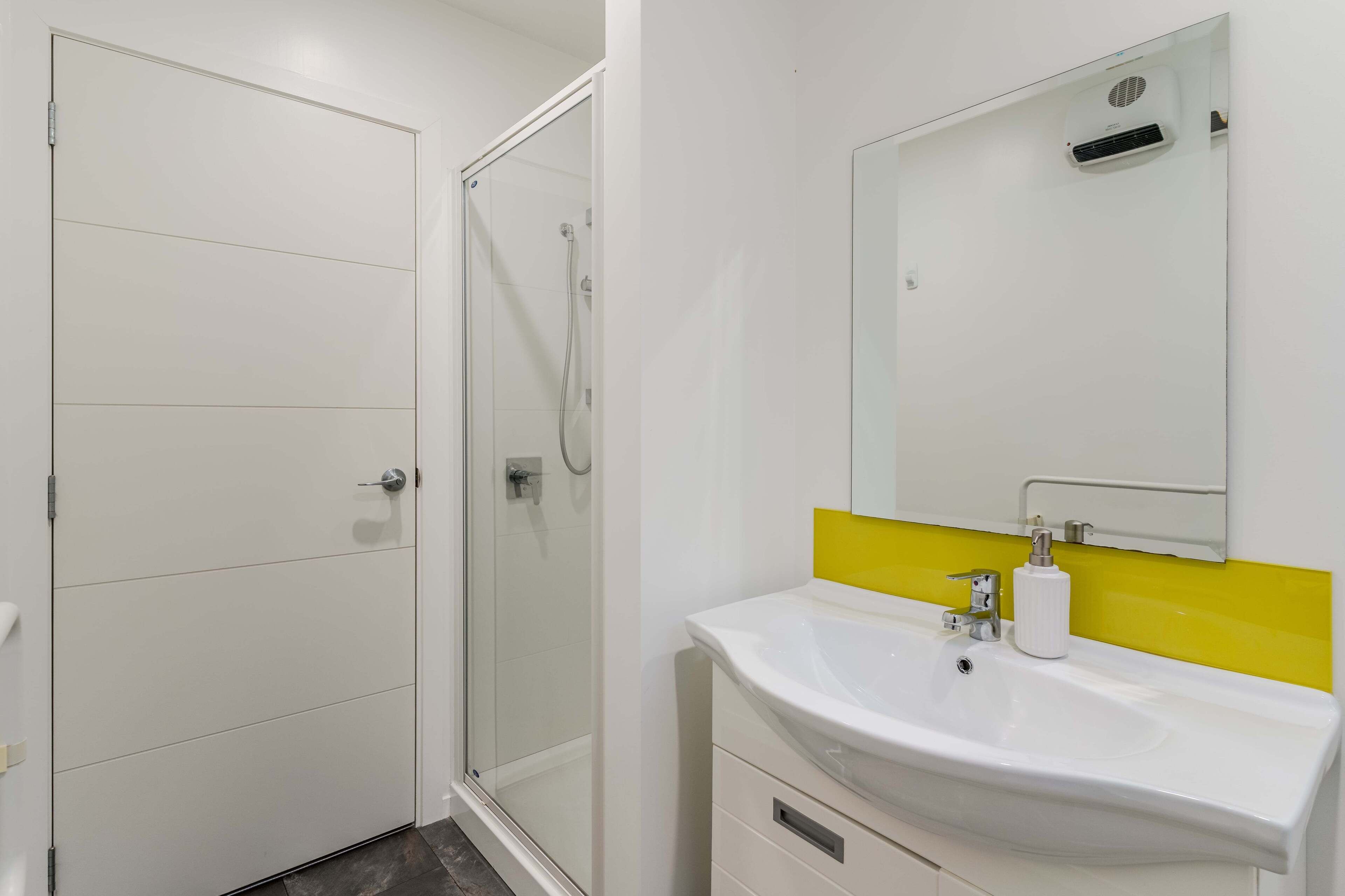 A modern bathroom with a glass shower enclosure, a white sink with a yellow accent backsplash, and a large mirror above the sink.