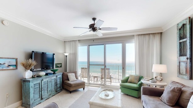 A spacious living room with a large window overlooking the sea, a ceiling fan, a television, and a green armchair. - Exclusive 2BR beachfront condo with gulf views (Miramar Beach)
