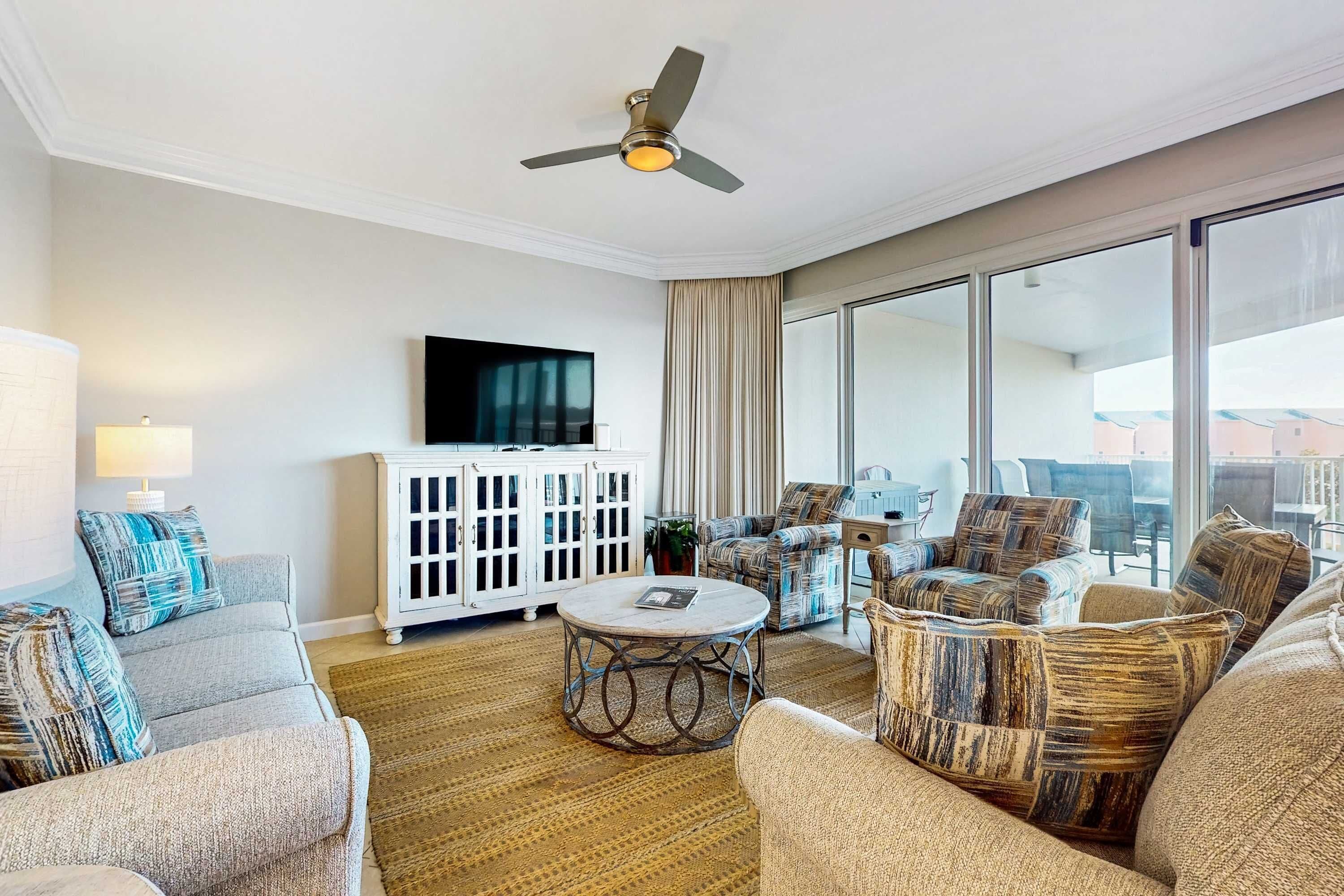 A spacious living room with a ceiling fan, a television mounted on the wall, and a large window offering a view of the city.