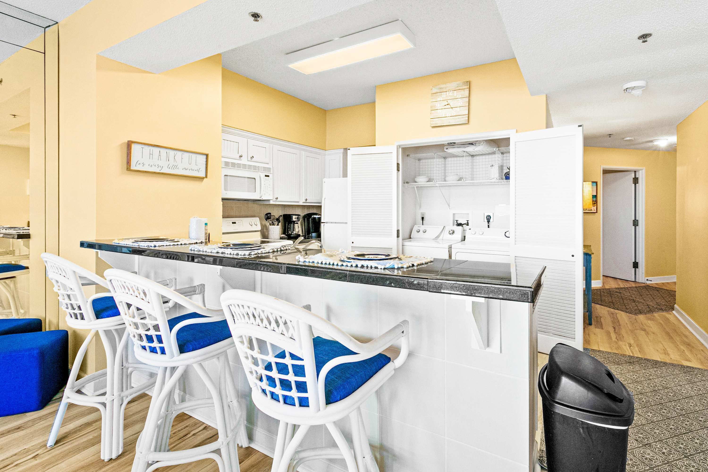A kitchen with white cabinets, a black countertop, and three white bar stools with blue cushions.