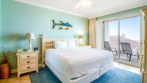 Un dormitorio con cama, mesitas de noche, ventilador de techo, un cuadro de peces y un balcón con sillas.