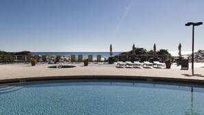 Zona de piscina con sillas de descanso y sombrillas, con vista despejada a la playa y un puesto de socorristas.