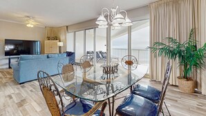 A spacious living room with a glass-top dining table surrounded by wicker chairs, a blue sofa, and a television. - Exclusive Gulf-Front Condo with Balcony (Miramar Beach)