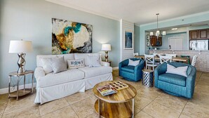 Ein Wohnzimmer mit einem weißen Sofa, blauen Sessel und einem Couchtisch, im Hintergrund eine Küche.