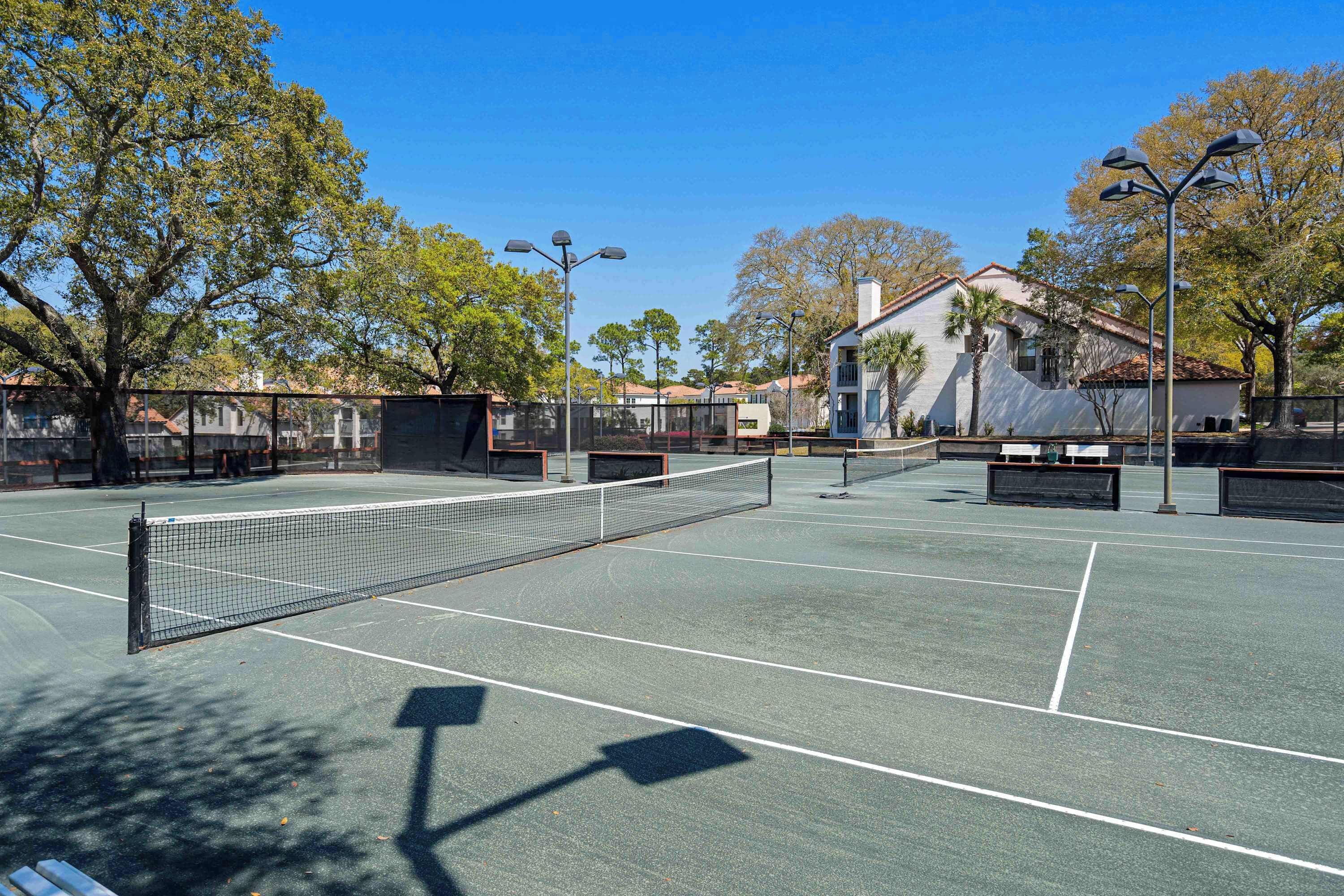 Un court de tennis en plein air, doté d’un filet, entouré d’arbres et de bâtiments.