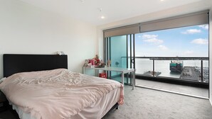 A modern hotel room with a large bed, a desk, and a balcony with a view of the water and a bridge.
