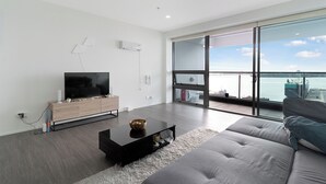 A modern living room with a large flat-screen TV, a wooden entertainment unit, a grey sofa, and a large window offering a view of the sea.