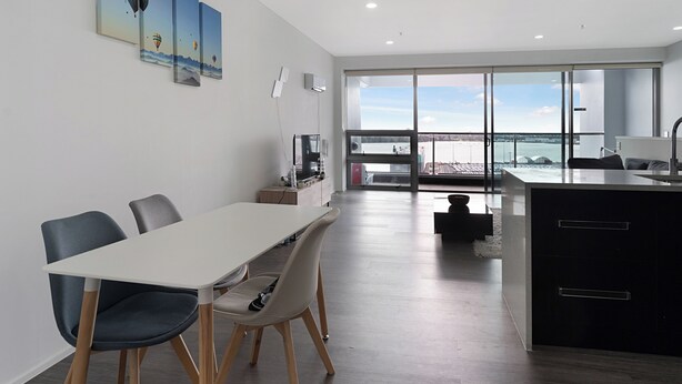 A modern dining area with a white table and chairs, a kitchen with a black countertop, and a large window offering a view of the sea.