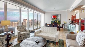A spacious living room with a large window, a white sofa, a round coffee table, and a dining area with a red pendant light. - Exclusive Gulf Views, Private Balcony (Miramar Beach)