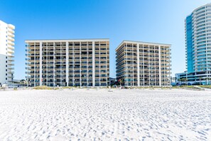 Ein Strandhotel mit mehreren Etagen, Balkonen und einem Sandstrand im Vordergrund.
