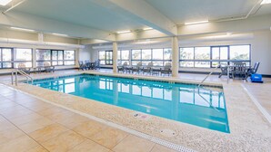 En indendørs swimmingpool med klart, blåt vand, omgivet af liggestole og borde, samt store vinduer der giver naturligt lys.
