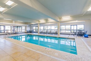 Ein Innenpool mit klarem, blauen Wasser, umgeben von Liegestühlen und Tischen, mit großen Fenstern für natürliches Licht.