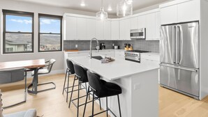 A modern kitchen with white cabinets, stainless steel appliances, a large island with bar stools, and a view of the mountains through the windows. - Mayflower Lakeside 104 | Reservoir + Mountain View | Full Kitchen | Private Bedrooms (Heber City)