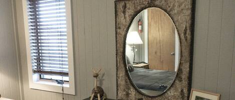 A bedroom with a dresser, a mirror, a window with blinds, a bed with a patterned blanket, and a small picture frame on the dresser.