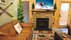 Un salon chaleureux avec une cheminée en pierre, un canapé en cuir, un sapin de Noël et un téléviseur diffusant une scène de ski.