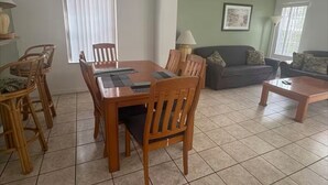 A dining area with a wooden table and chairs, a sofa, and a coffee table in a living room with tiled flooring and green walls. - Home in Kissimmee near Disney and Universal (Kissimmee)