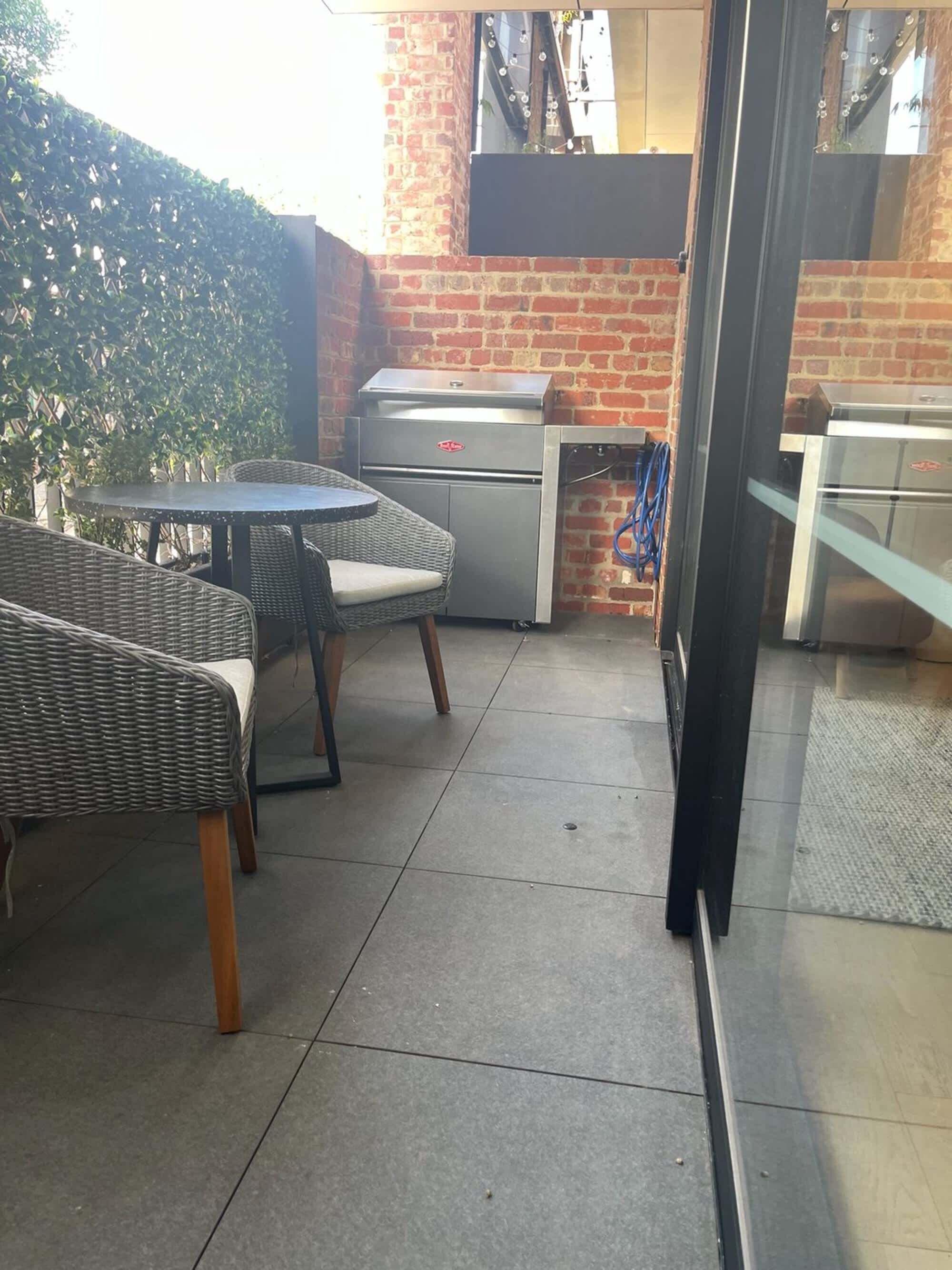 An outdoor patio with wicker furniture, a grill, and a glass door leading to a brick building.