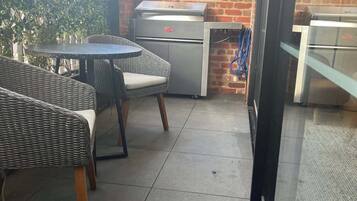 An outdoor patio with wicker furniture, a grill, and a glass door leading to a brick building.