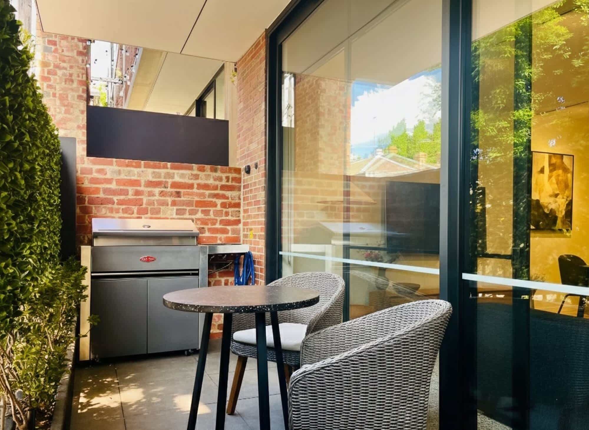 A patio area with a grill, wicker chairs, and a glass wall offering a view of the outdoors.